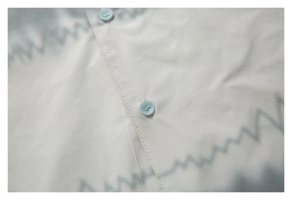 Close-up of pale gray shirt fabric, a button placket, blue-gray buttons, and part of a dark jagged pattern.