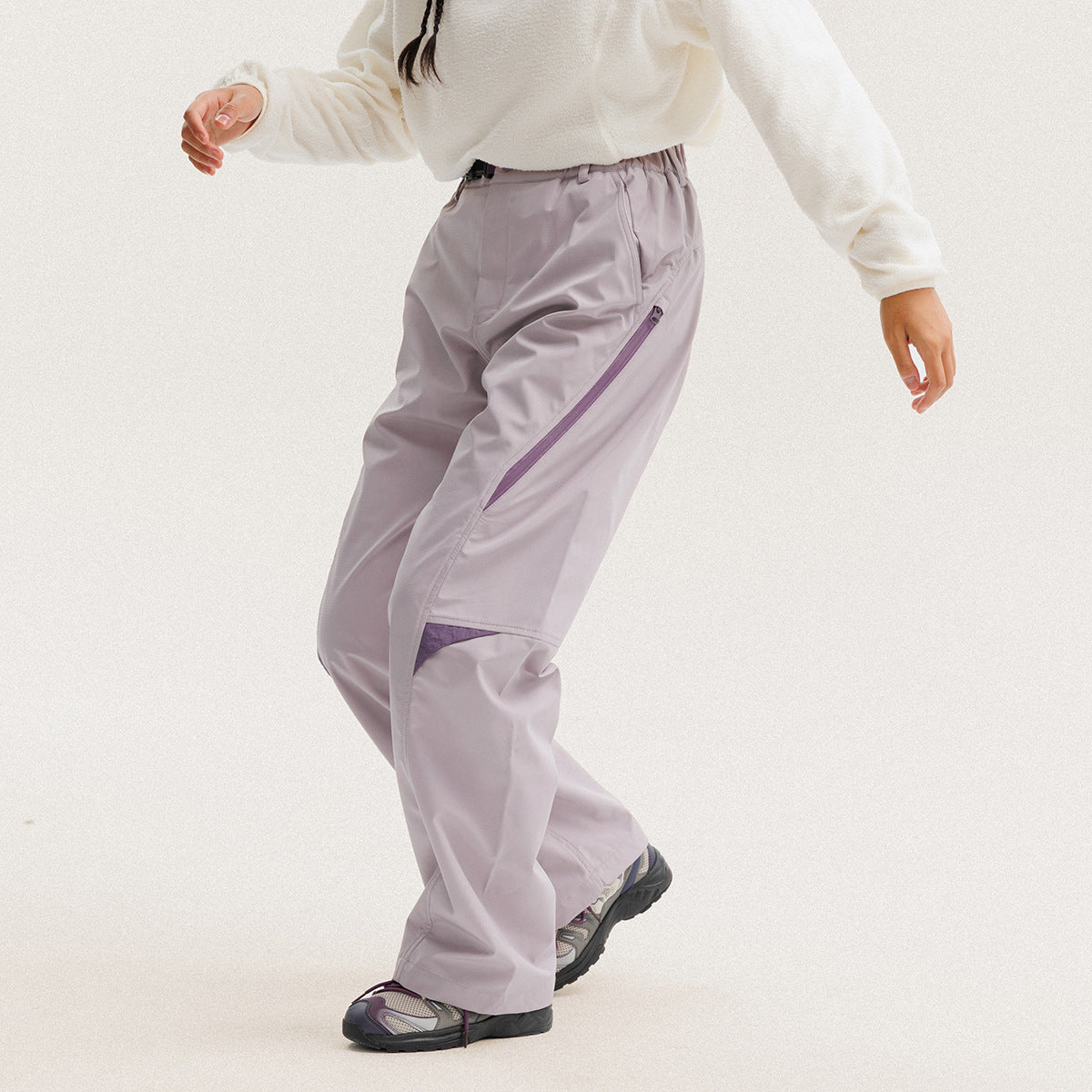 Model wearing loose pale lavender trousers with a purple diagonal zip pocket and contrasting knee panel.