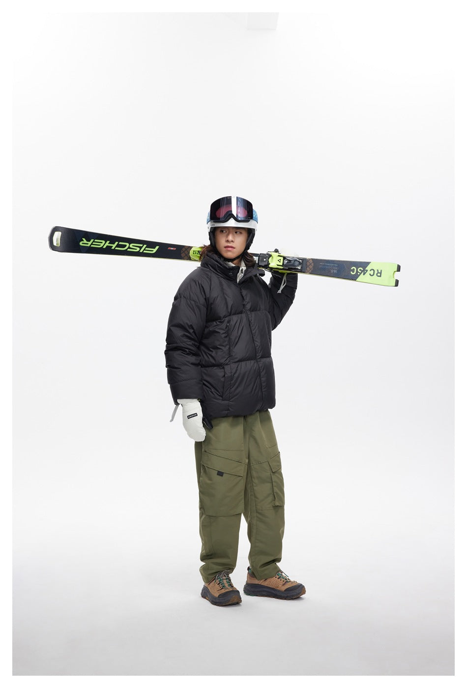 Model wearing a black puffer jacket and olive cargo pants, holding skis across one shoulder.