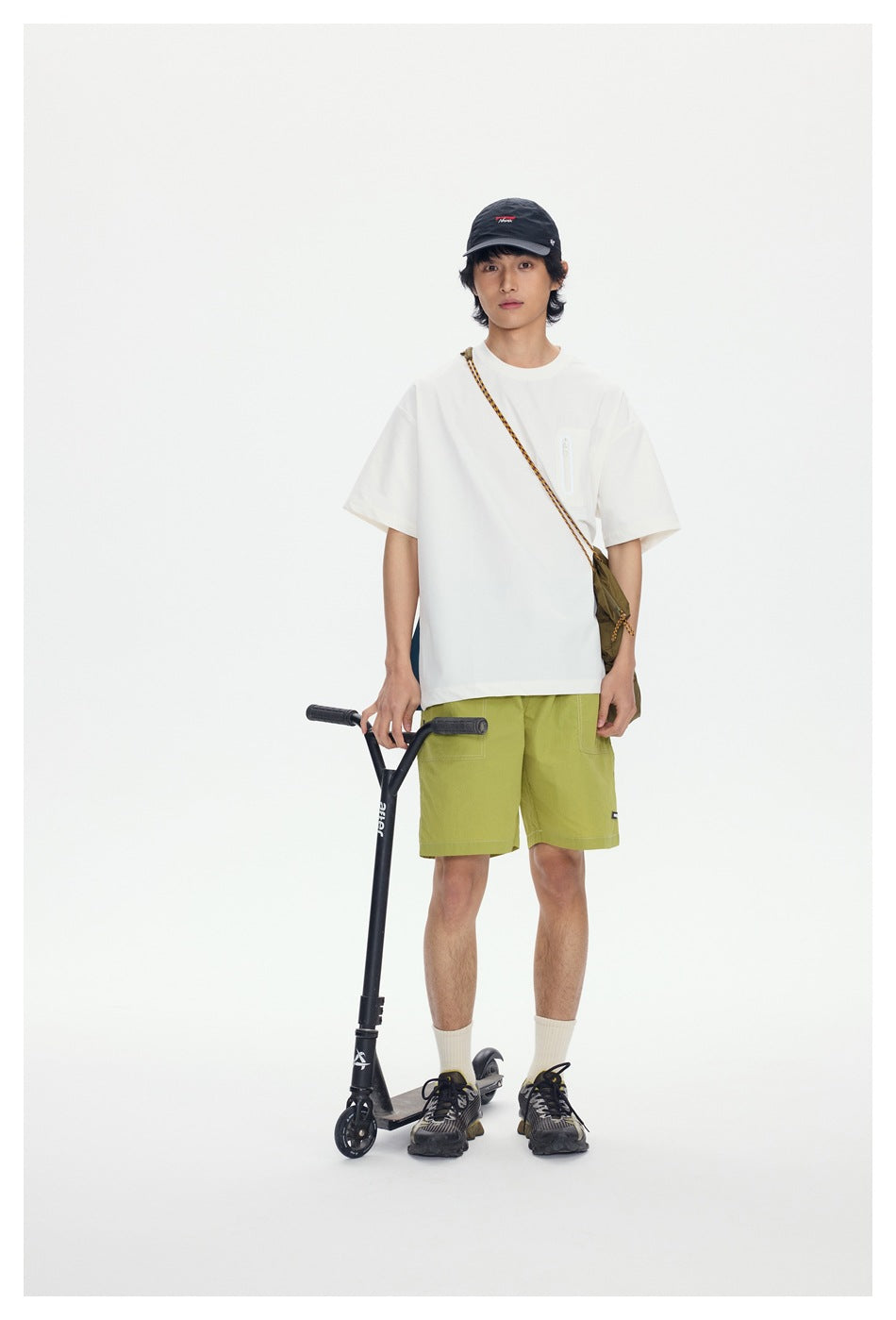 Model wearing a white T-shirt and lime-green shorts, standing beside a scooter.