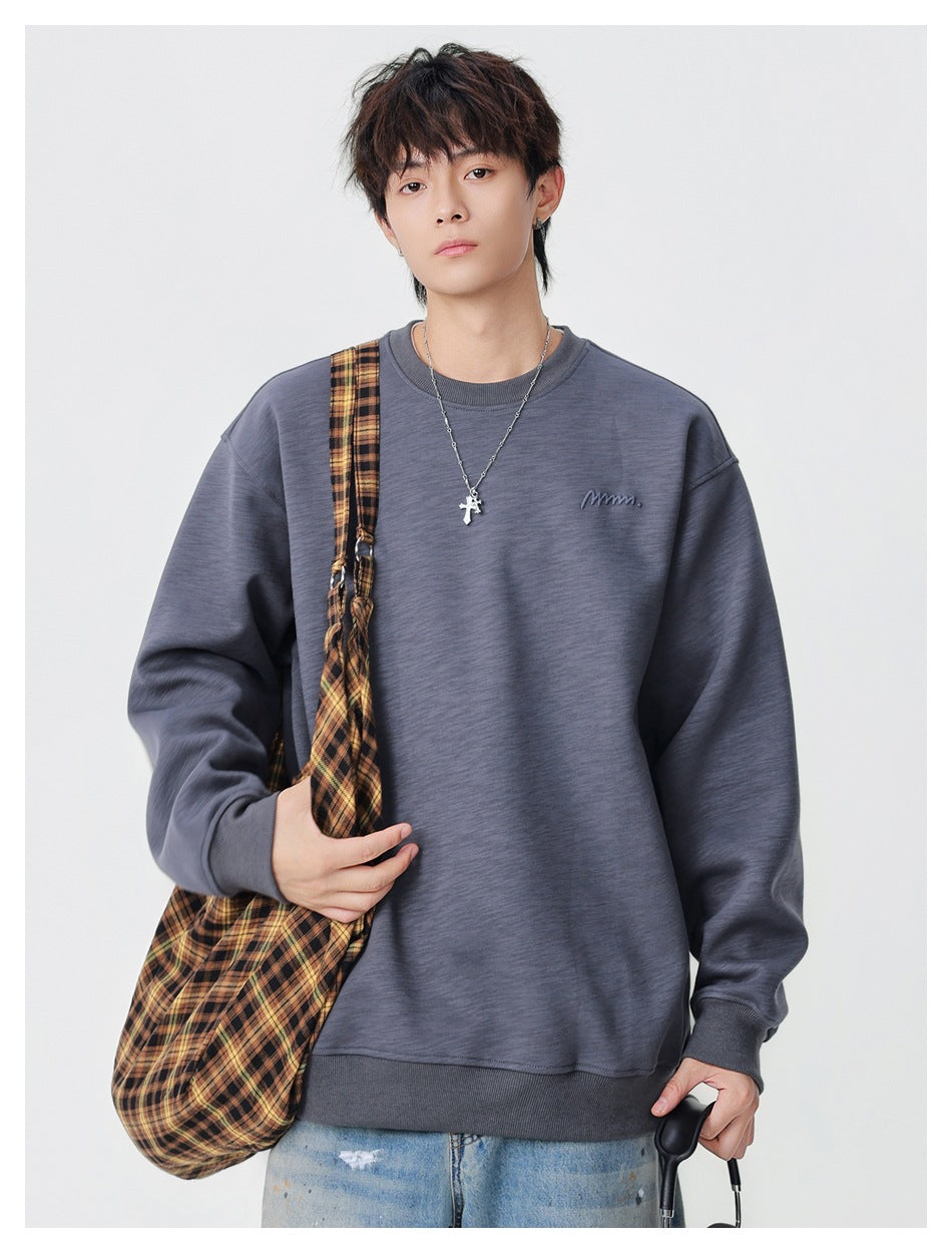 Model wearing a charcoal gray oversized crewneck sweatshirt with a small embroidered chest mark, carrying a plaid shoulder