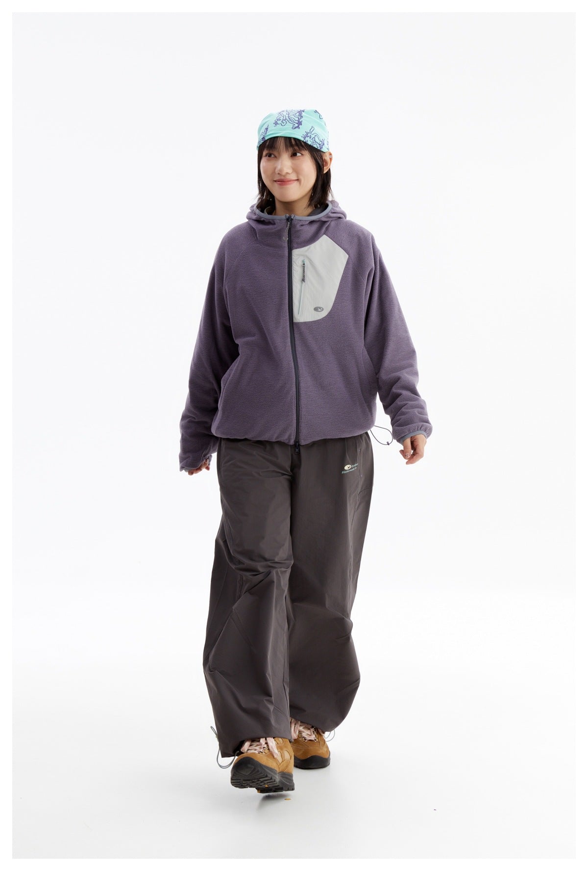 Model walking in a purple hooded zip jacket with a pale chest pocket, dark wide-leg pants and hiking shoes.