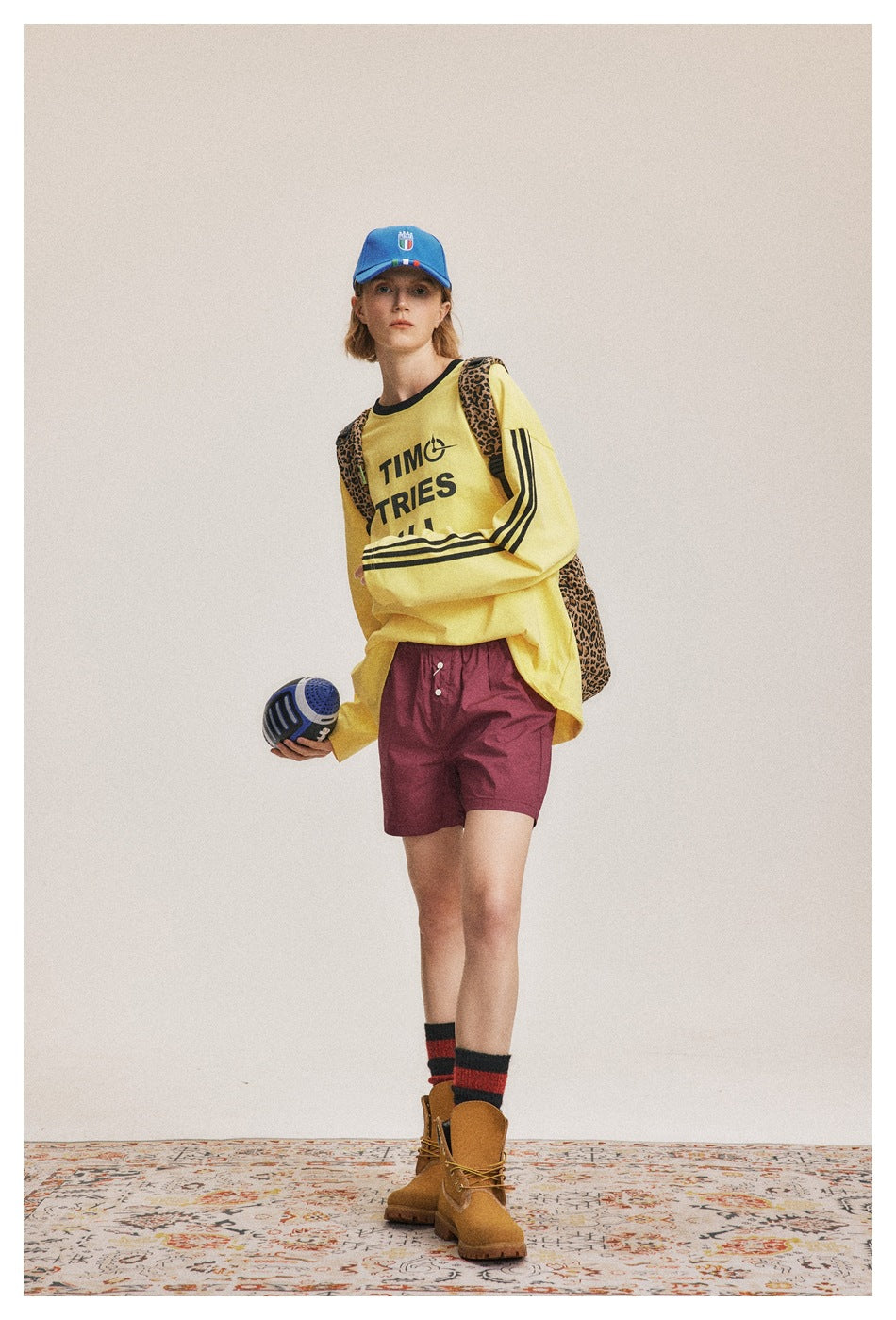 Model facing front wearing the yellow long-sleeve top with black sleeve stripes, burgundy shorts and a blue cap