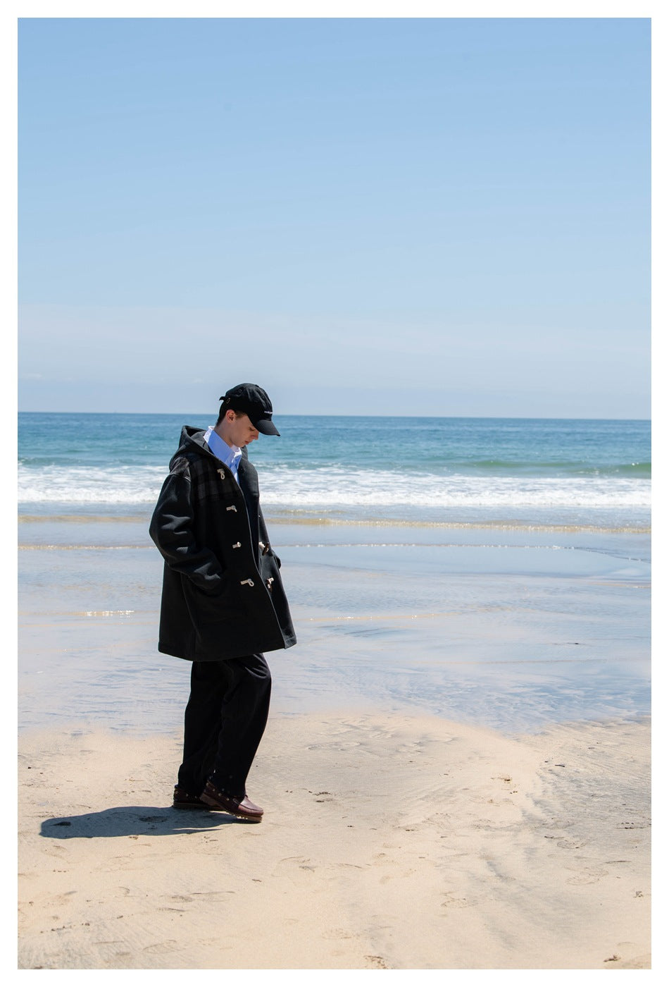 Full-length angled view of a person wearing a dark hooded duffle coat on the beach.
