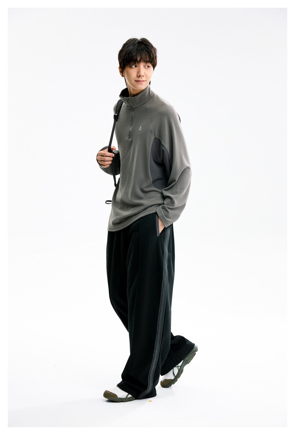 Model wearing a gray quarter-zip pullover with black side panels and loose black trousers with a side stripe.