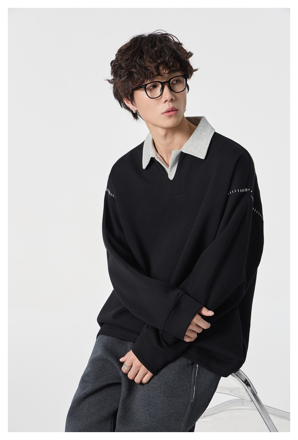 Model wearing the black pullover with gray collar and white shoulder stitching, seated with arms crossed, gray pants