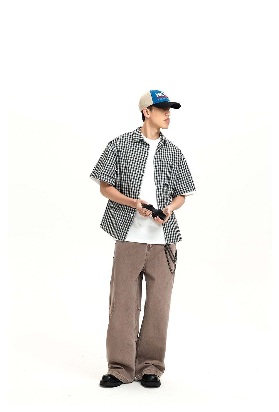 Man in wide-leg brown jeans with a checked shirt, white tee and cap, shown at a slight angle with a wallet chain