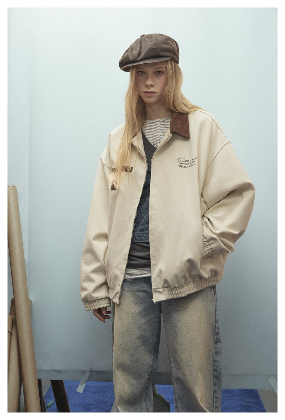 Model wearing a cream zip jacket with brown collar and chest details, paired with faded jeans and a brown cap.