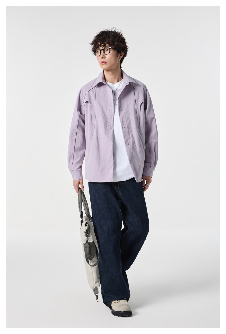 Model wearing an open pale lavender shirt over a white tee with dark wide-leg jeans, carrying a light shoulder bag.