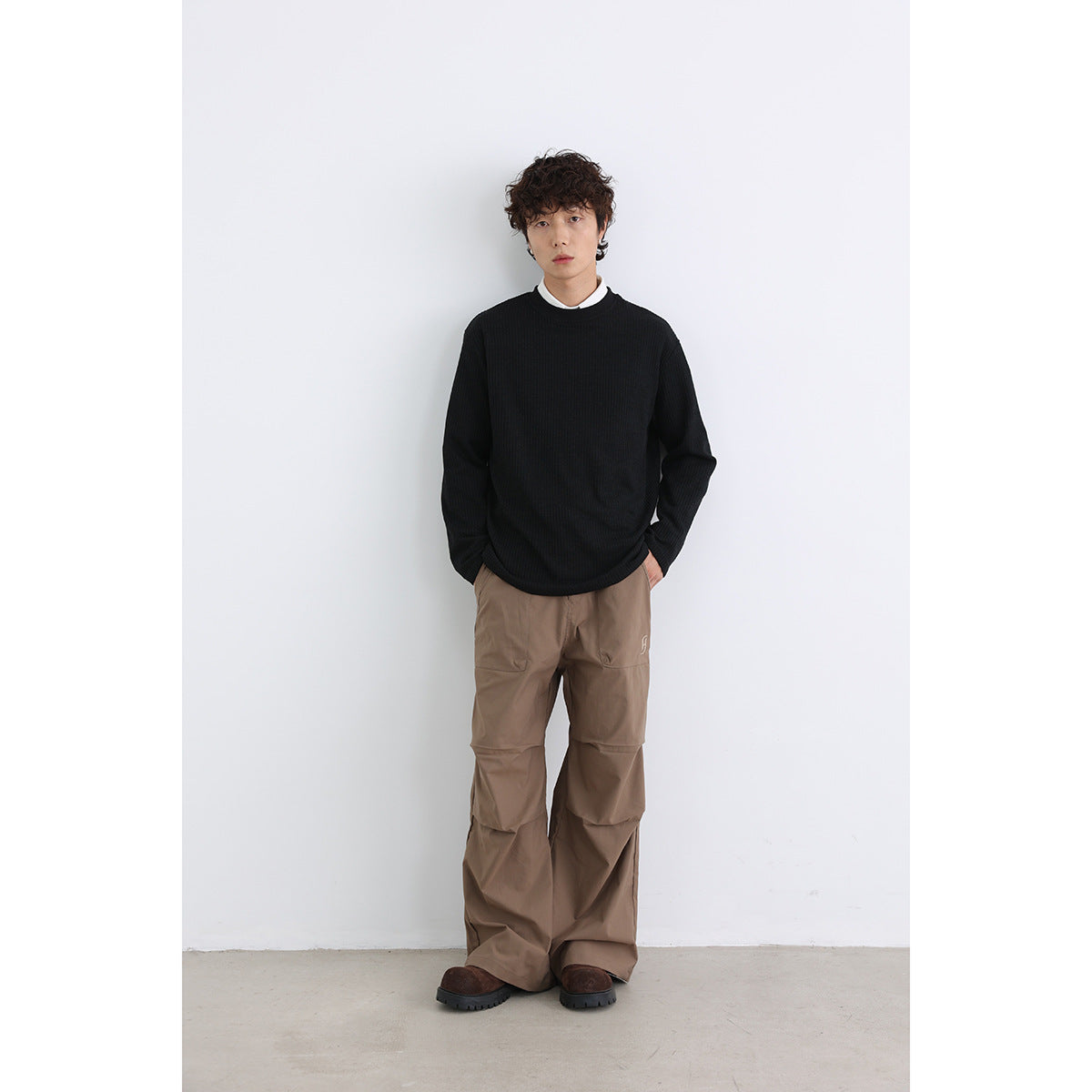 Model wearing a black ribbed crewneck sweater over a white collar, with loose brown trousers.