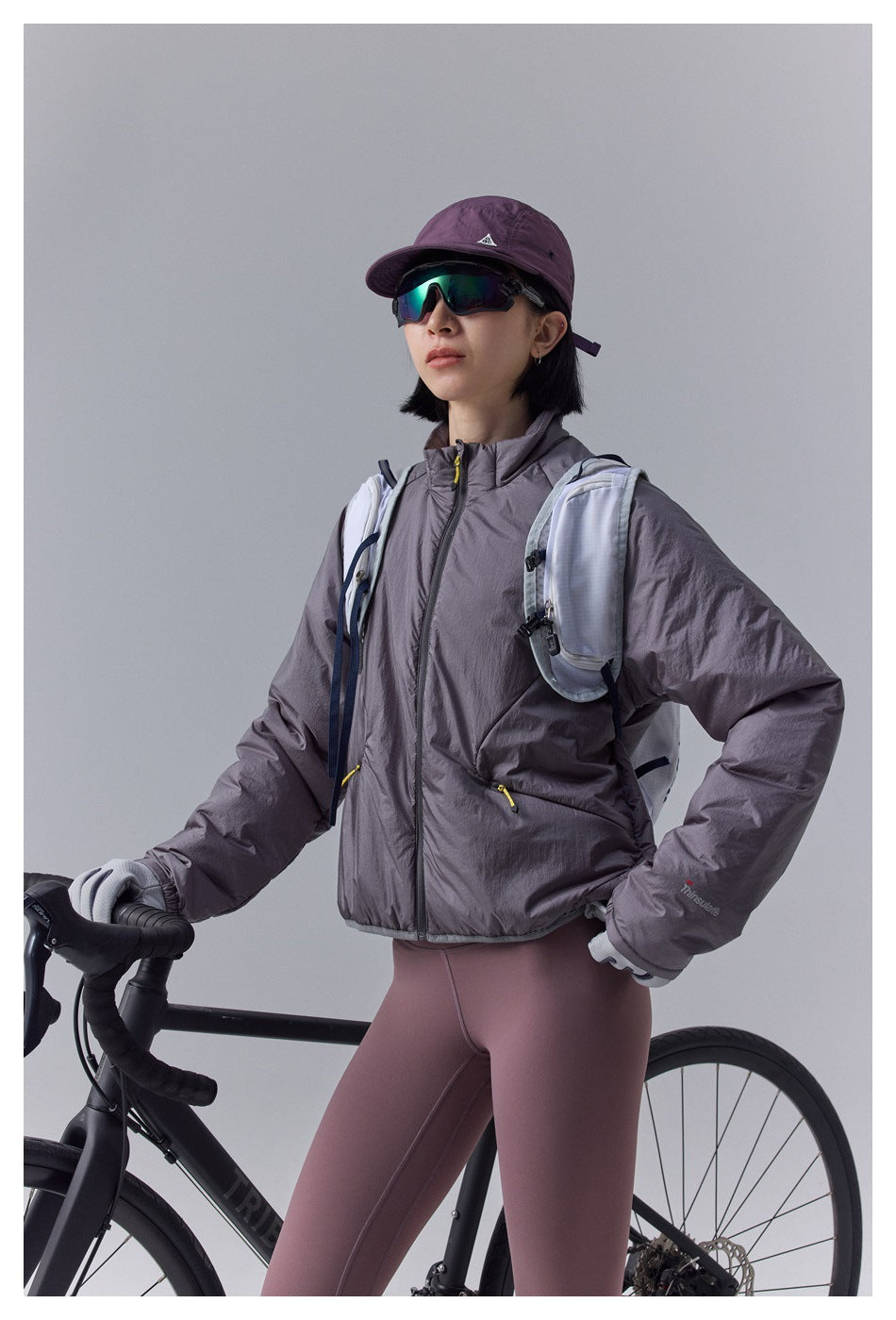 Model wearing a gray-plum padded jacket and mauve leggings beside a road bike, shown from the front in a close view.
