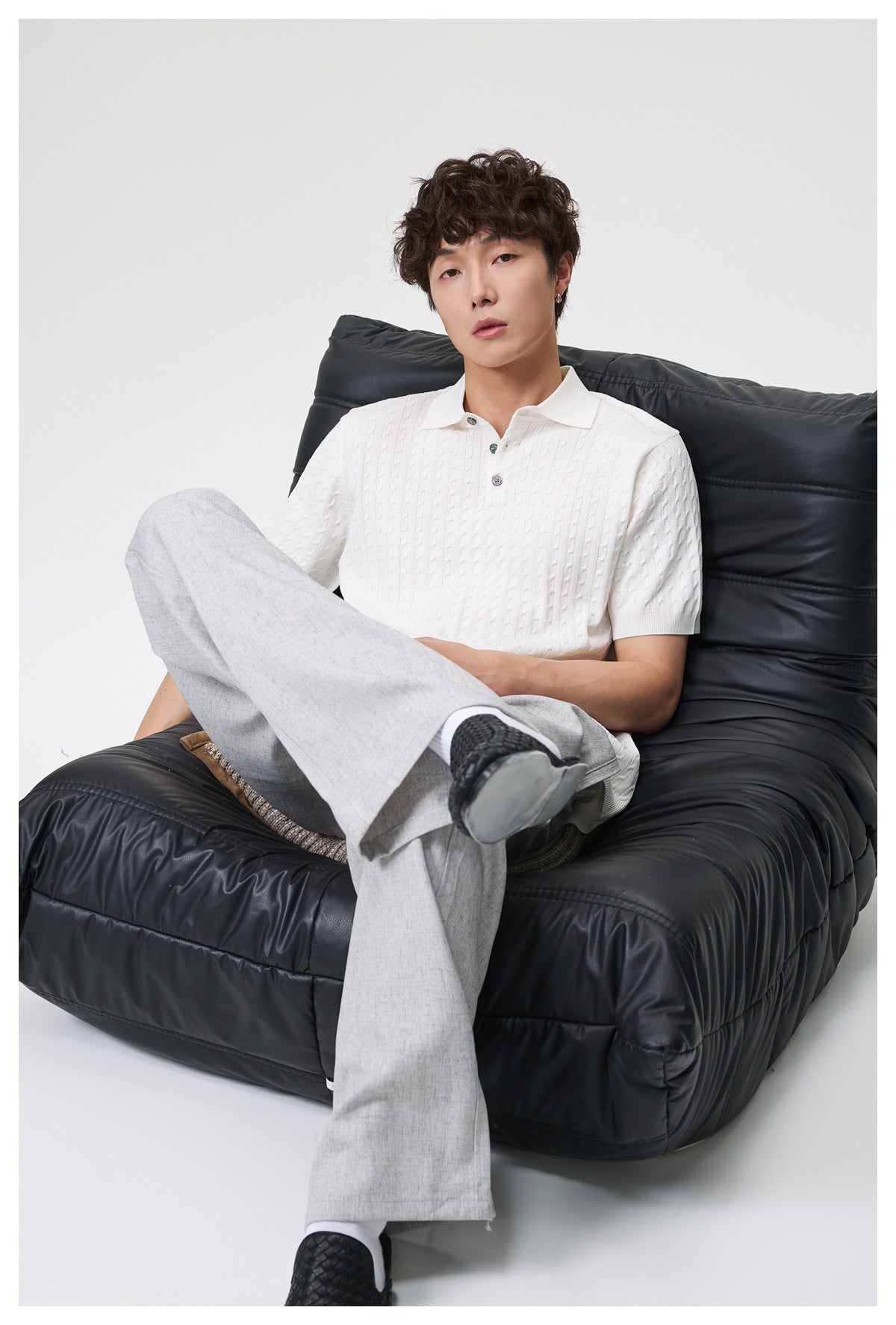 Model seated in a black chair, wearing a white short-sleeve cable-knit polo and light gray trousers.