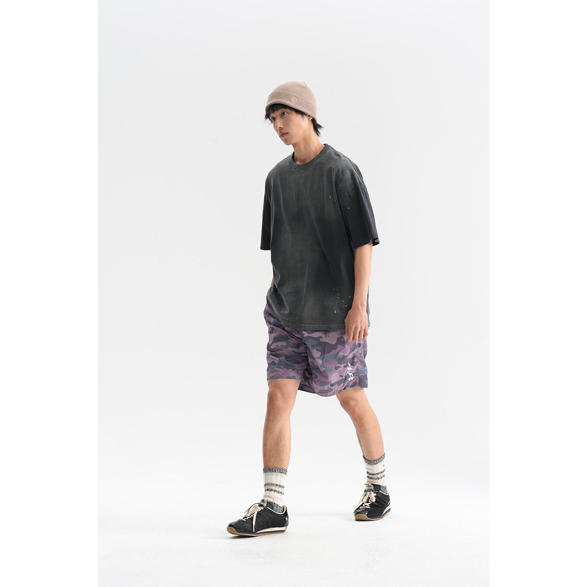 Model wearing a charcoal oversized T-shirt, purple camouflage shorts, and a beige knit beanie, shown at an angle.