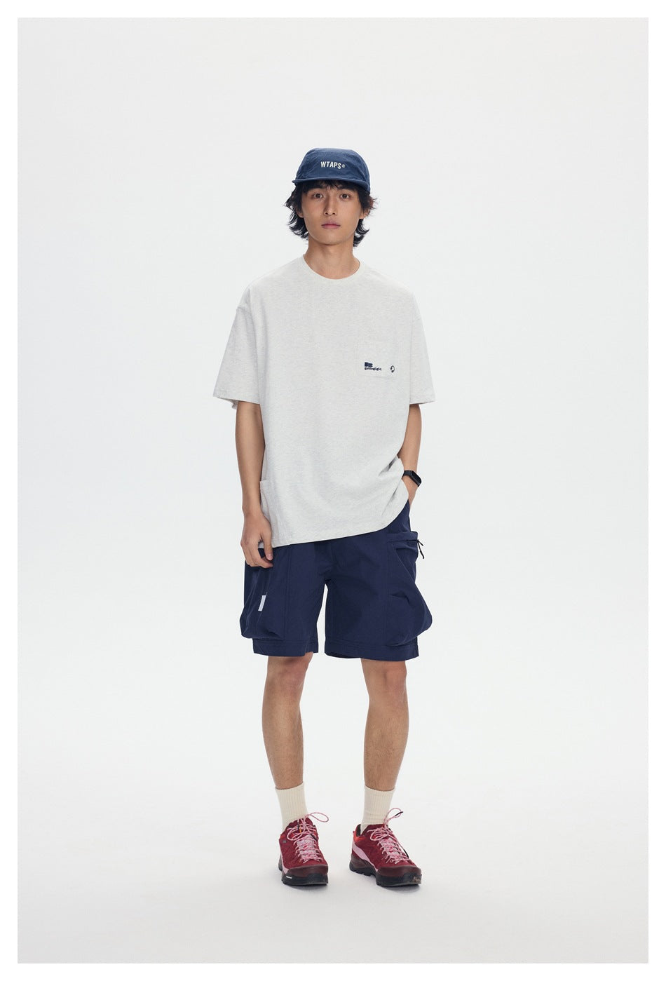Model wearing navy cargo shorts, a light gray T-shirt, navy cap, cream socks, and red hiking shoes; front view.