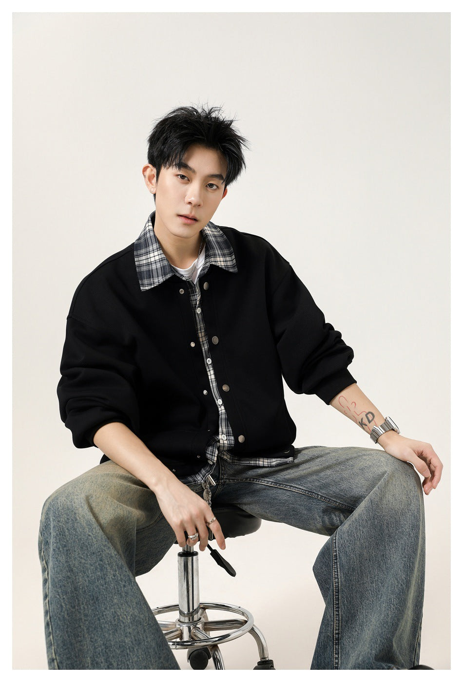 Seated model wearing an oversized black snap-front jacket over a gray plaid shirt and white tee.