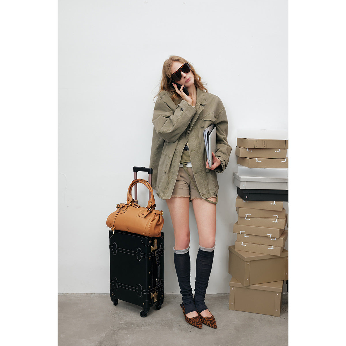 Model wearing an oversized olive utility jacket with large pockets and a button front, styled with shorts and dark leg