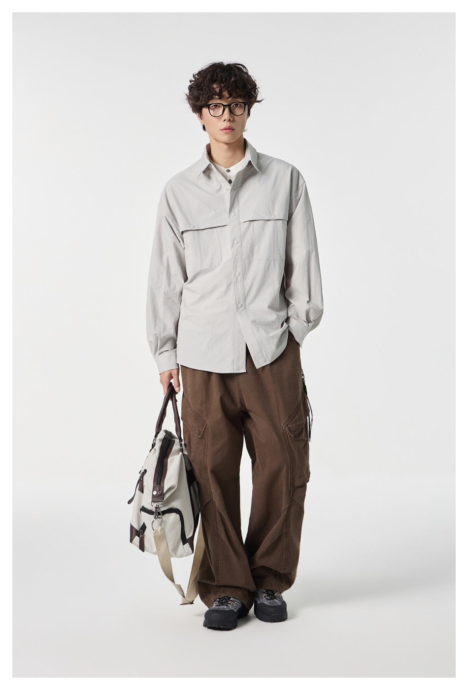 Model wearing a light gray button-front shirt with flap chest pockets and brown cargo trousers, carrying a bag.