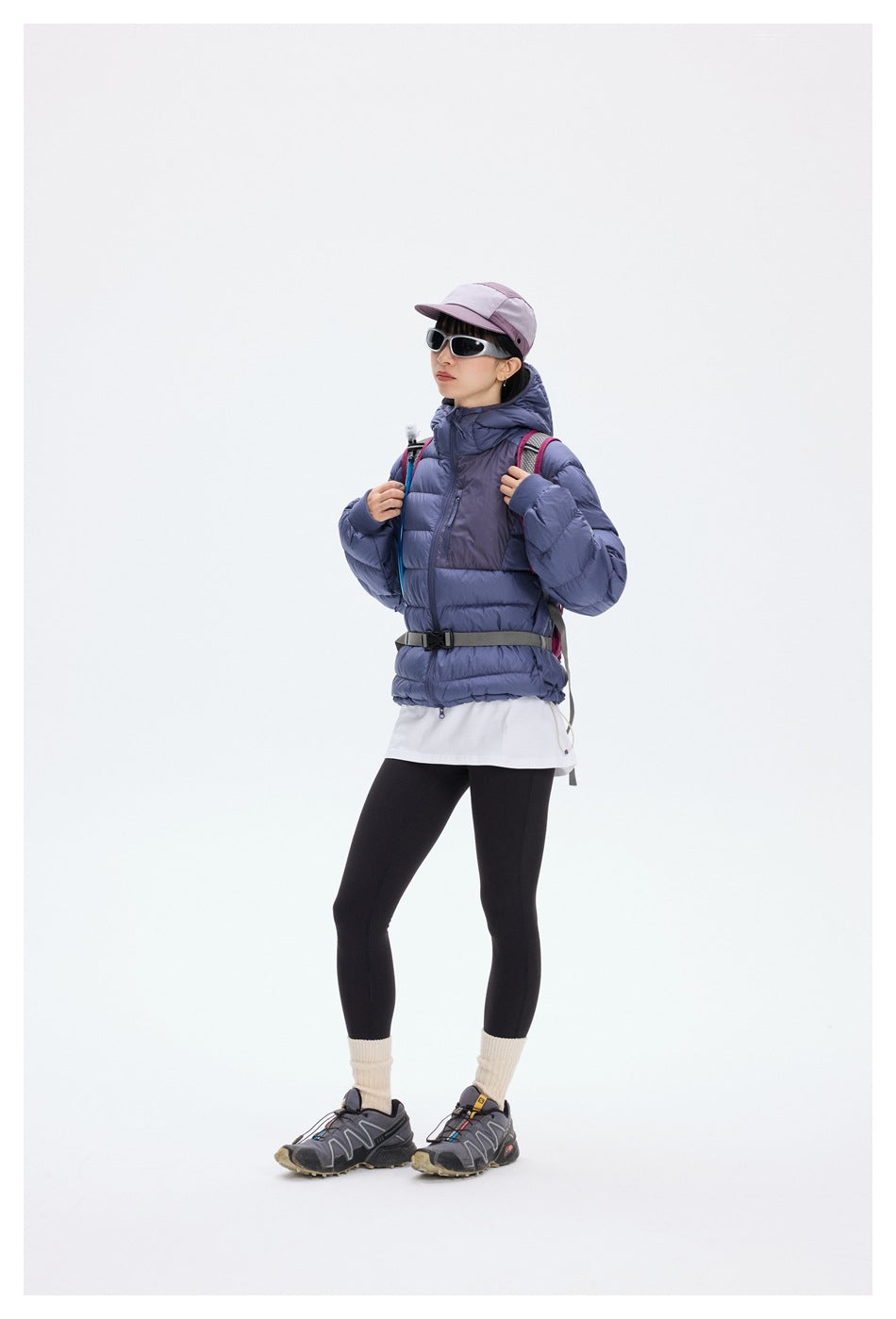 Model wearing purple hooded quilted jacket with cap, sunglasses, backpack, black leggings and trail shoes, full length