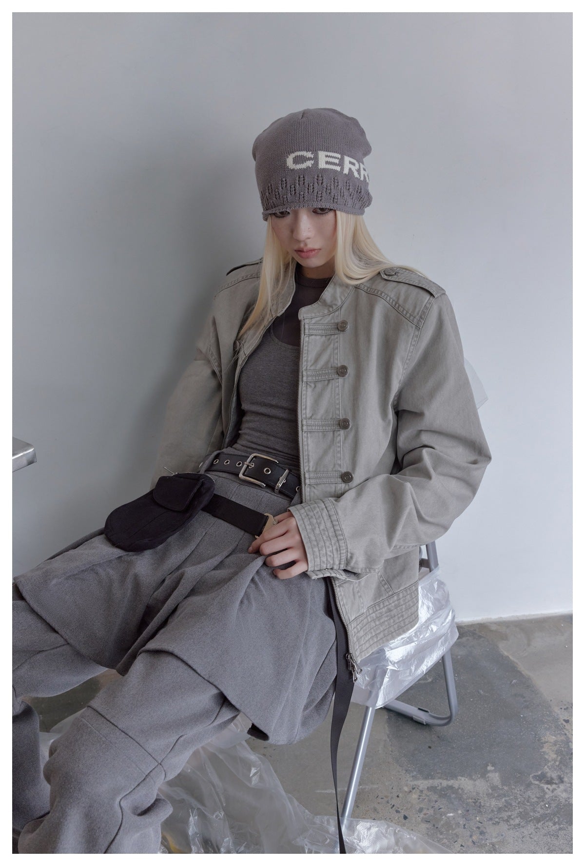 Seated model in a light gray military-style jacket with button tabs, zipper, and shoulder epaulettes.