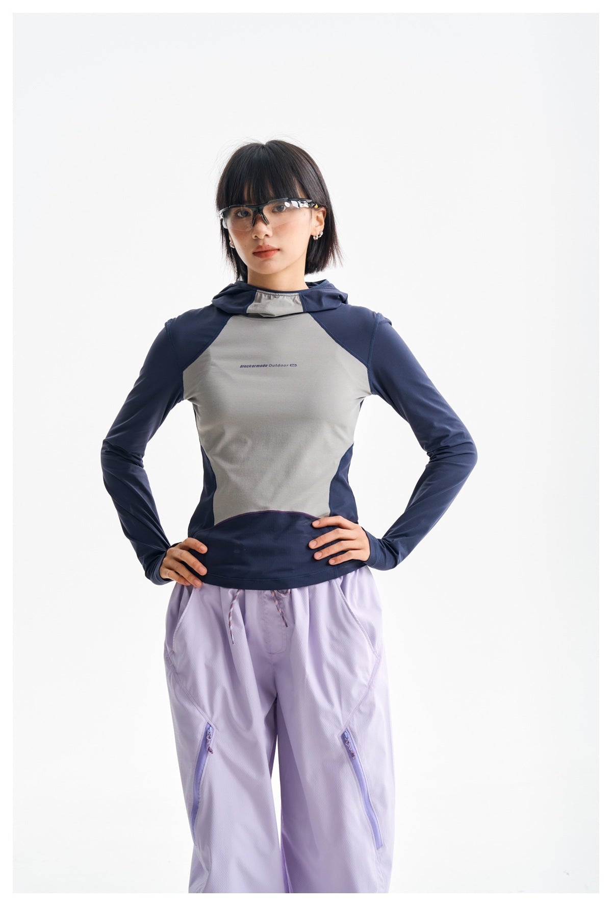 Model wearing a color-block navy and gray hooded top with lavender pants, hands on hips.