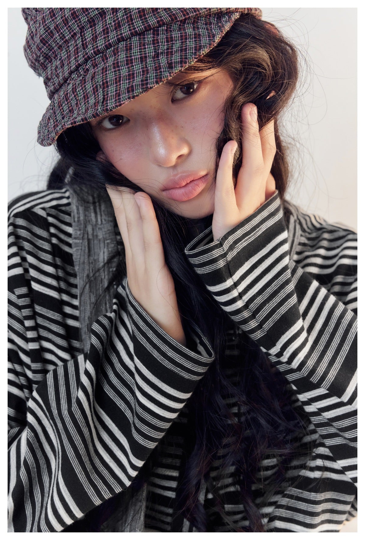 Close-up of a model in a plaid cap with hands at face, showing the black and white striped long sleeves