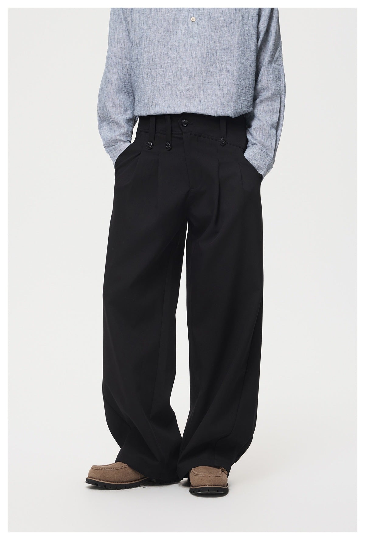 Front view of a model wearing black wide-leg trousers with a buttoned, tabbed waistband and brown shoes.