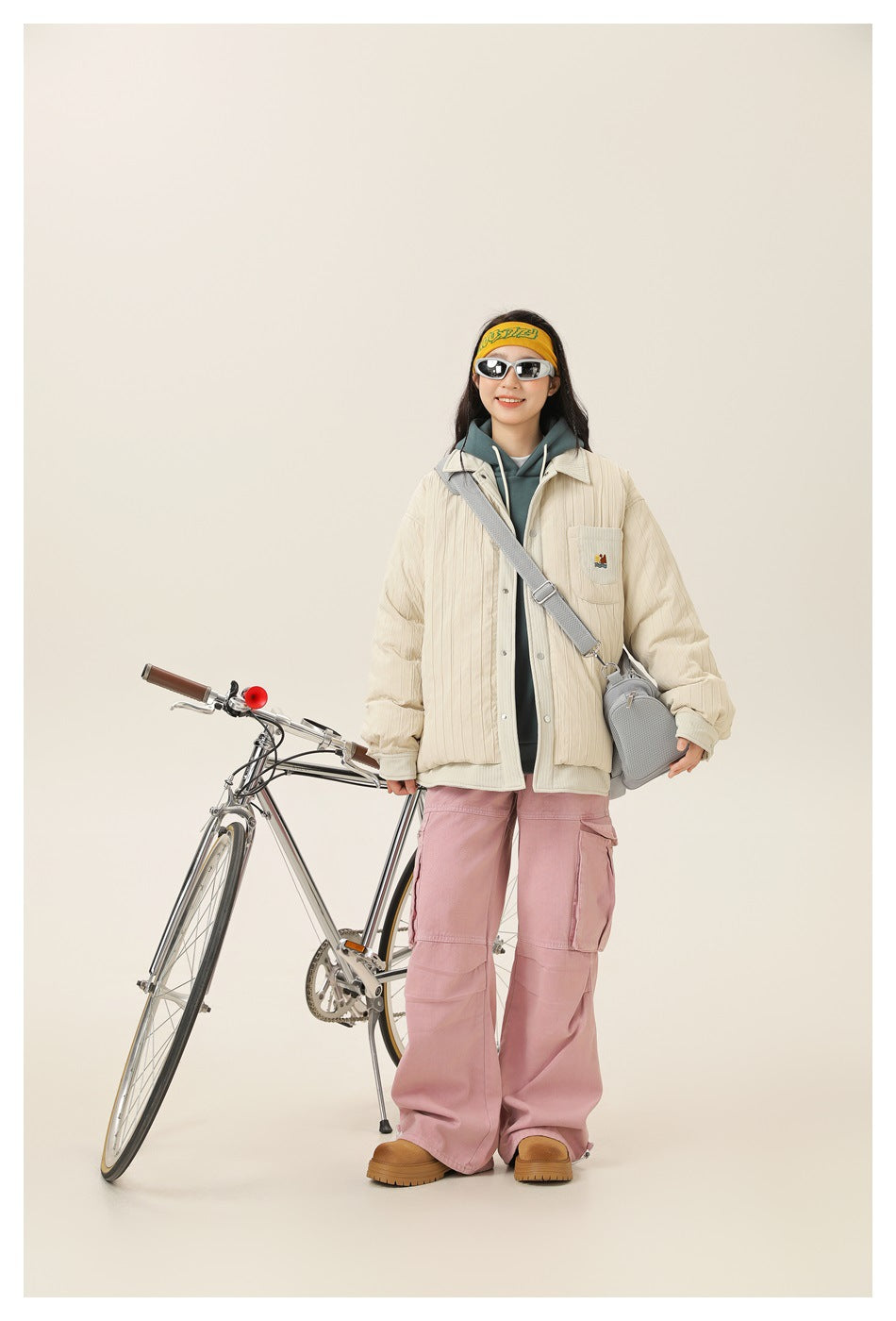 Full-length front view of a model in a cream quilted jacket and pink cargo pants beside a bicycle.