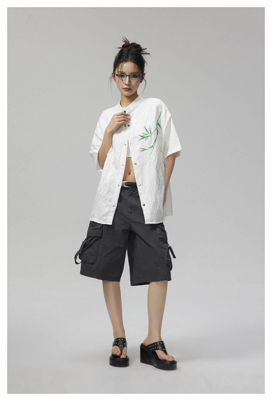 Model wearing a white short-sleeve shirt with green bamboo embroidery, dark cargo shorts, and platform sandals.