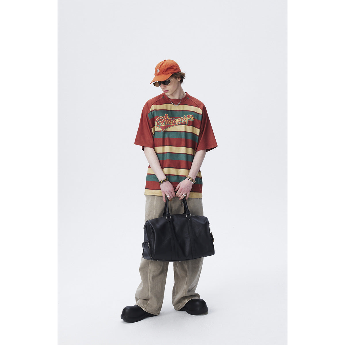 Model wearing a rust-red, teal, and beige striped oversized T-shirt with chest lettering, beige trousers, and black shoes.