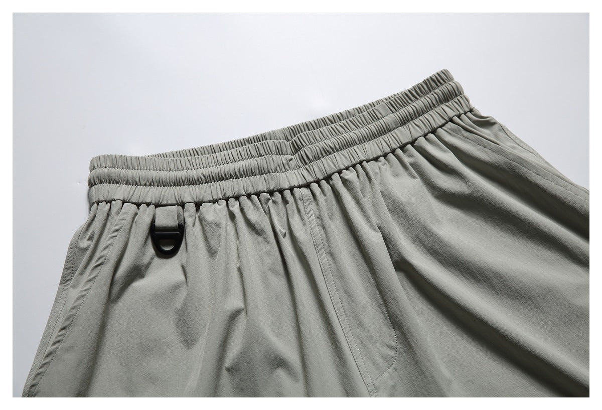 Close-up of gray-green shorts showing the gathered elastic waistband, black D-ring, and front pocket seam.