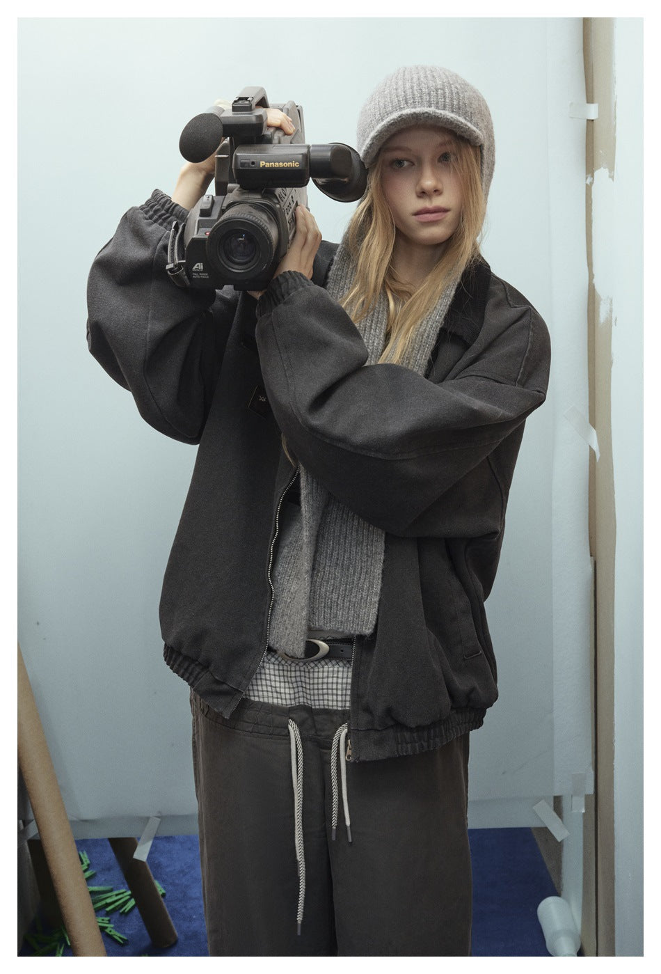 Model wearing an oversized charcoal jacket, gray knit cap and scarf, and dark pants while holding a video camera.