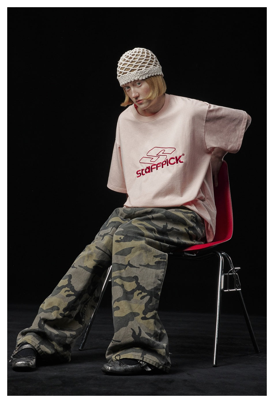 Seated model wearing a pale pink graphic T-shirt, loose camouflage trousers, and a cream open-knit hat.