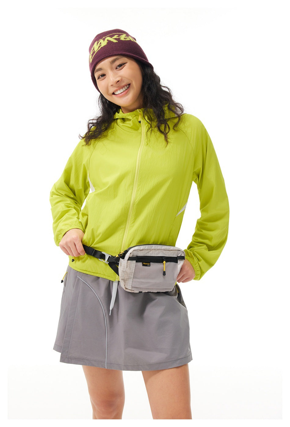 Model wearing a lime hooded jacket, gray skirt, waist pack, and burgundy beanie with yellow lettering.