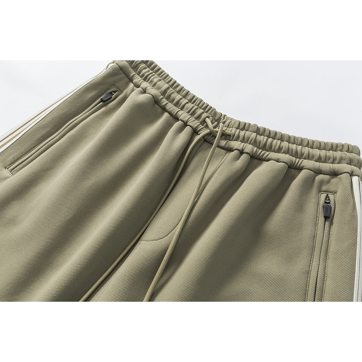 Close-up of olive jogger pants showing the elastic drawstring waist, zip pockets and white side stripe.