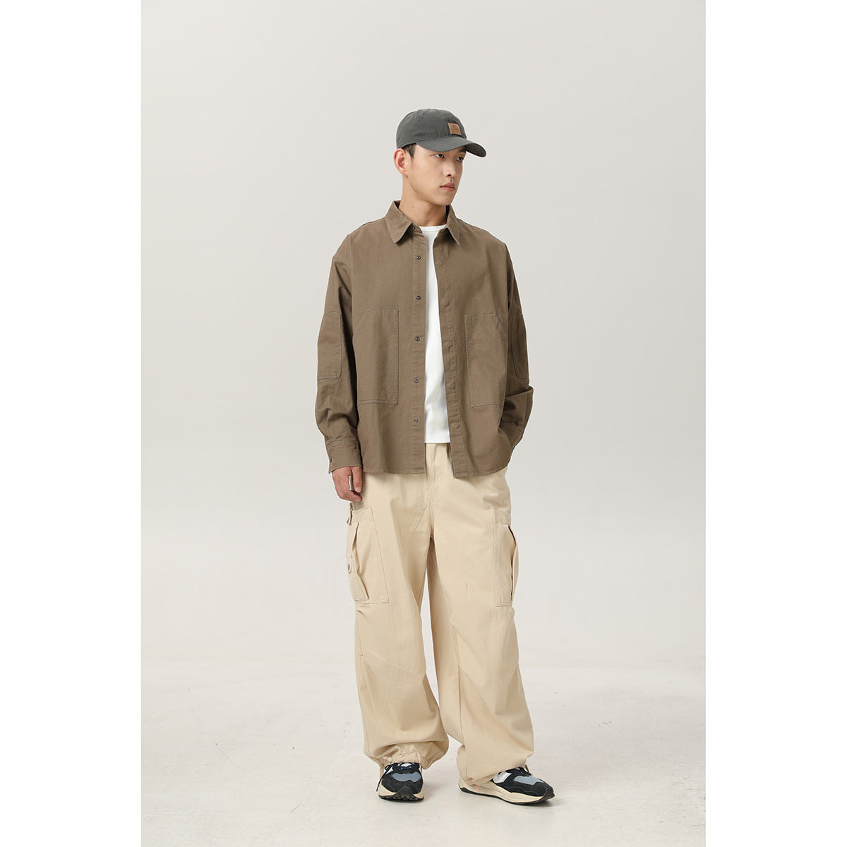 Model wearing a brown button-front overshirt with large pockets, cream cargo pants, a dark cap and sneakers.
