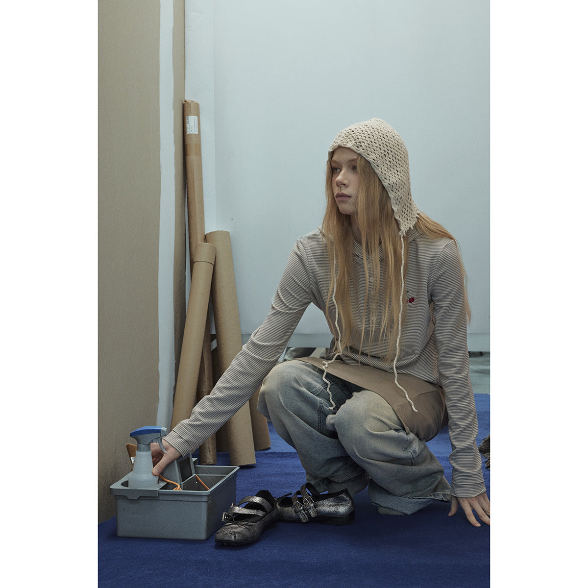 Model crouching in a light gray striped hooded top and loose jeans, wearing a cream crocheted hood.