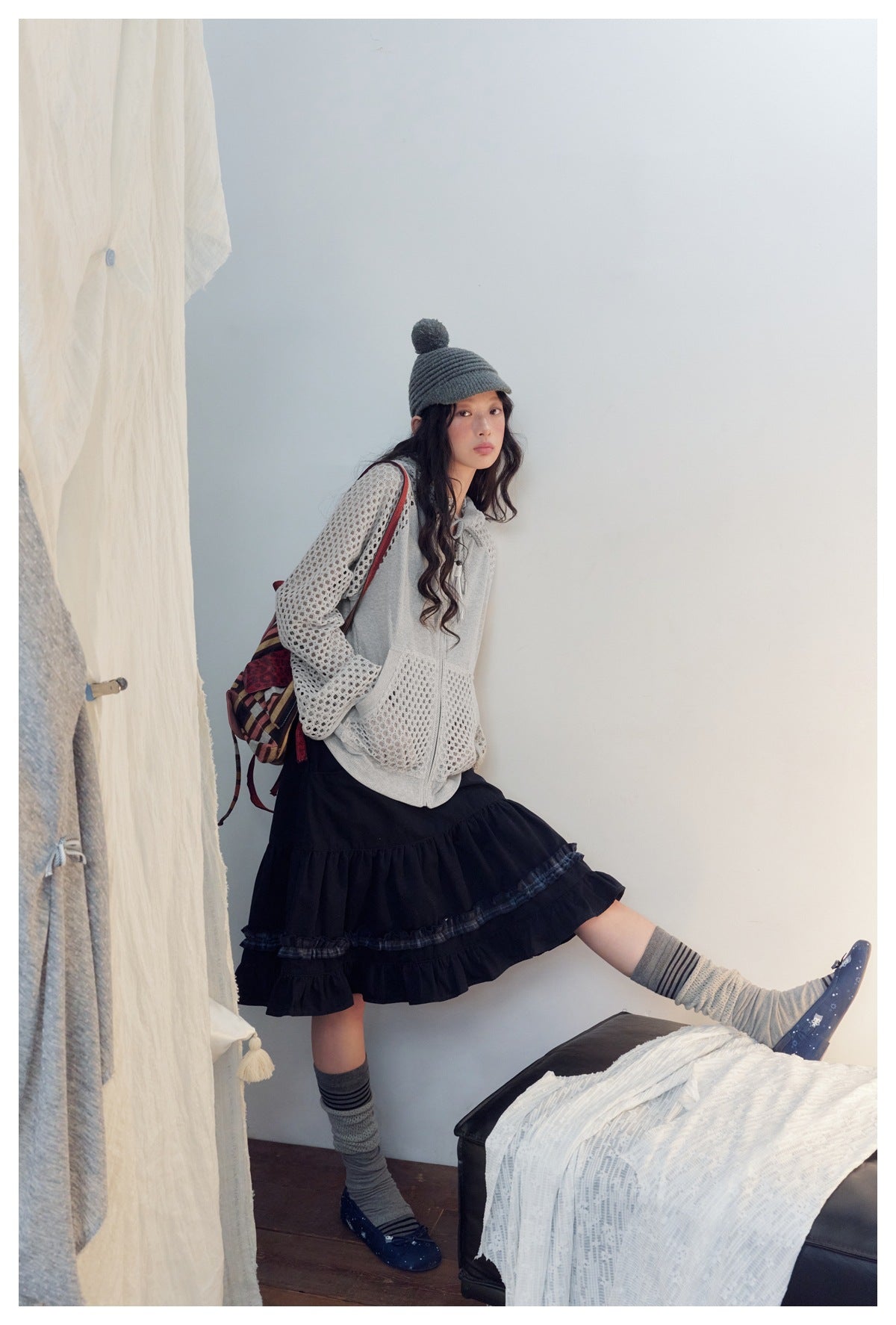Full-length view of a model wearing a light gray mesh hoodie, black ruffled skirt, gray leg warmers, and flats.
