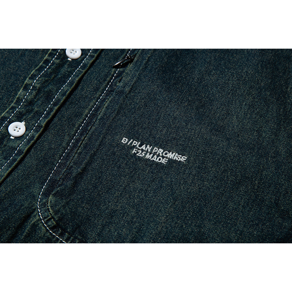 Close-up of a dark blue-green shirt front with white buttons, a curved pocket edge, and small white embroidery.
