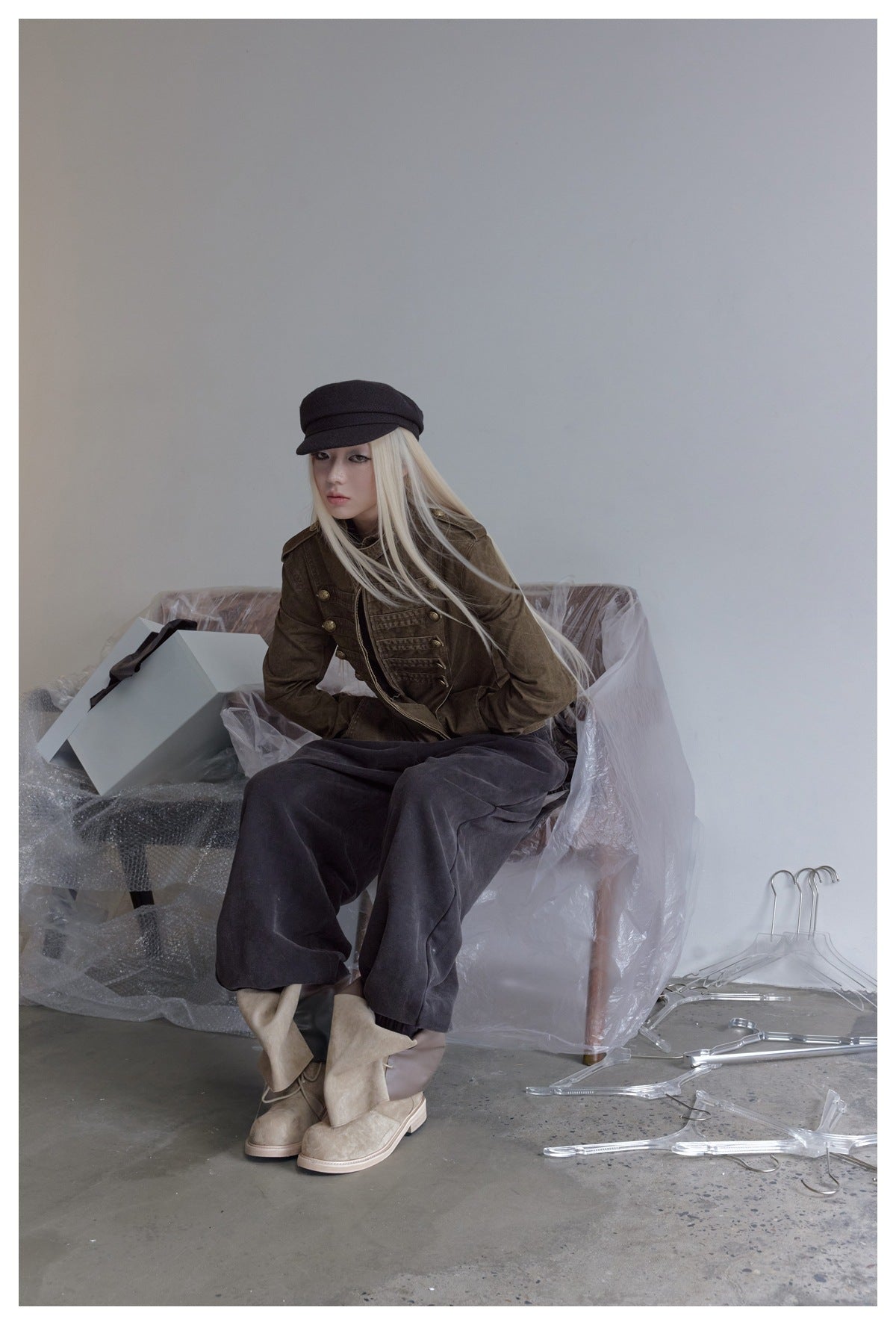 Seated model wearing a dark olive-brown buttoned jacket, charcoal trousers, and a black cap.