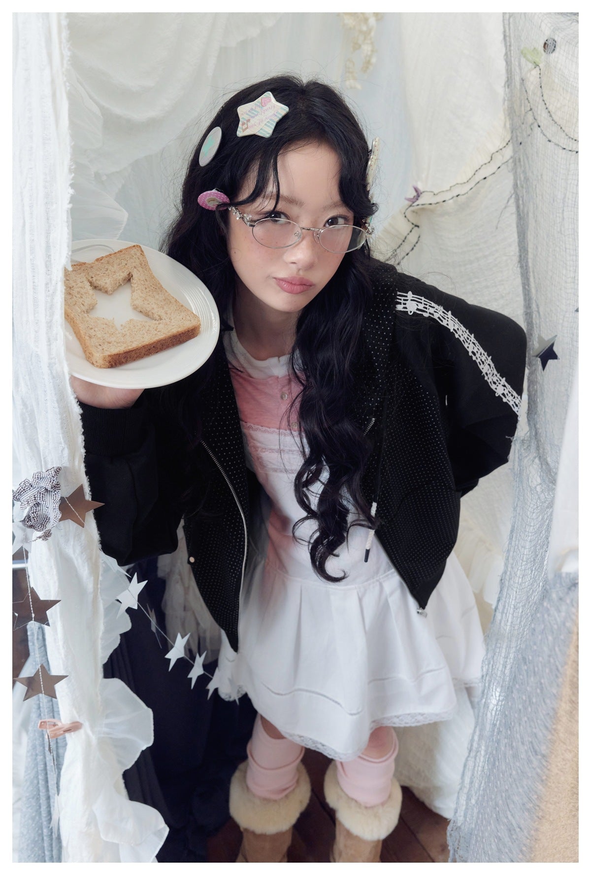 Model in a black dotted zip hoodie with white music-note sleeve trim and a pink-and-white dress, holding a plate.