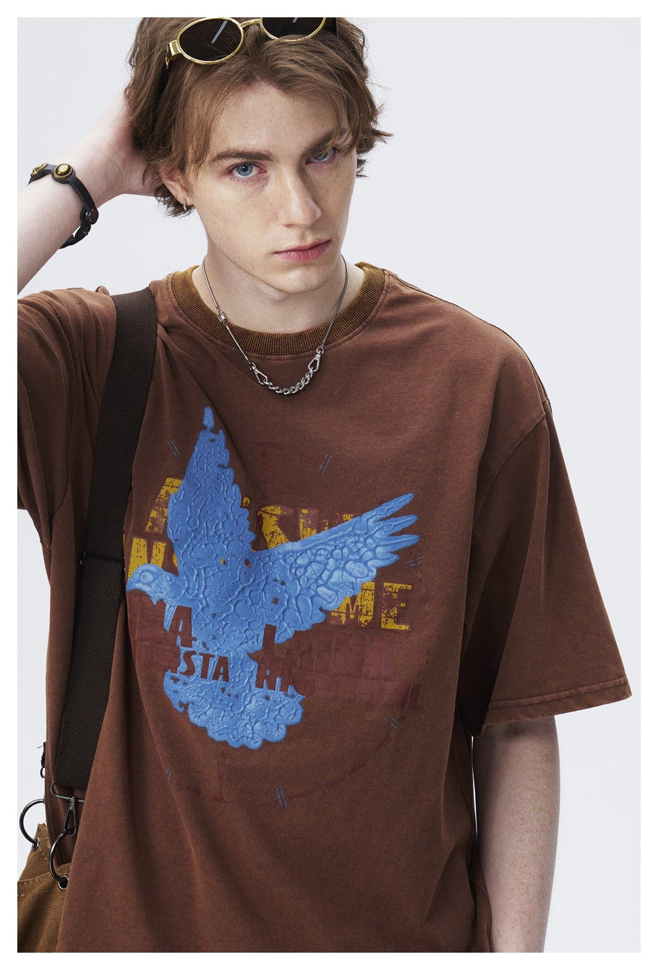 Model wearing an oversized brown T-shirt with a large raised blue bird graphic and distressed lettering.