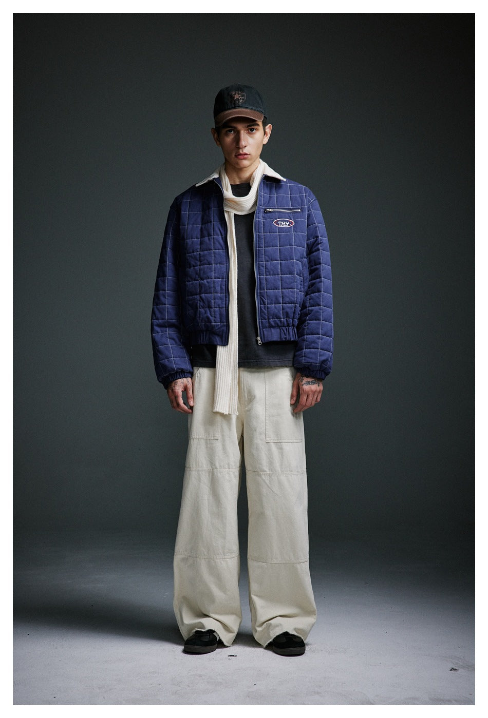 Man wearing the blue quilted zip jacket over a dark tee with a cream scarf, cream wide-leg pants and a cap
