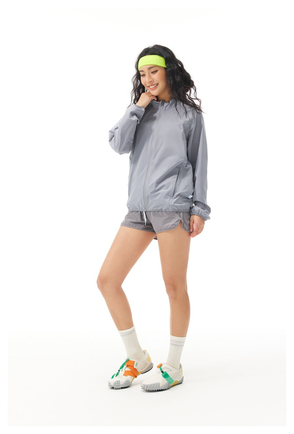 Angled view of a model in a light gray zip jacket and matching shorts with a neon yellow headband.