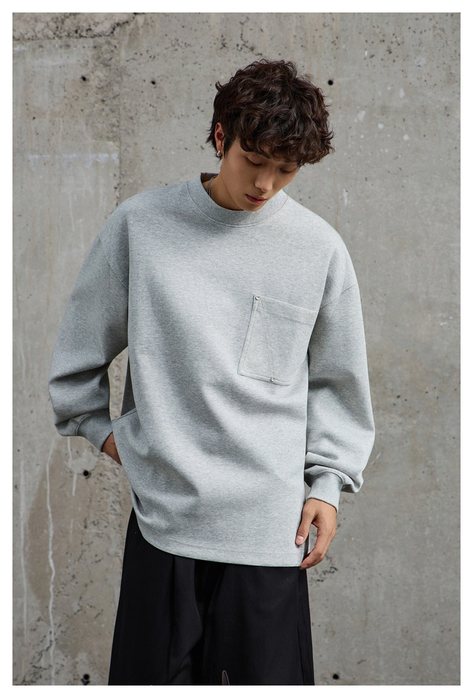 Model wearing a gray oversized crew neck sweatshirt with a chest pocket and black pants against a concrete wall