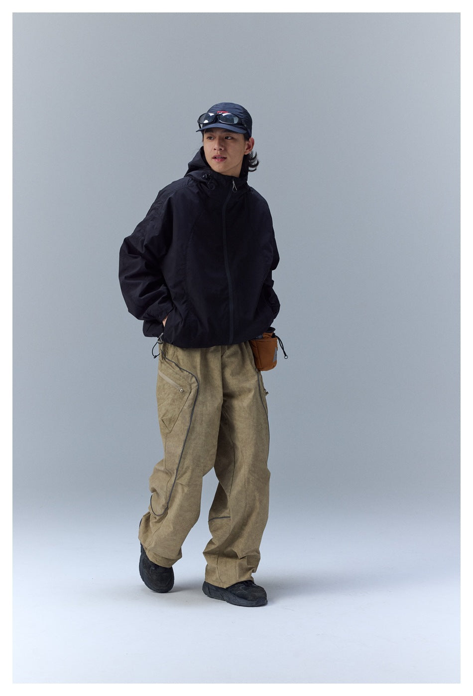 Model wearing a black hooded jacket and loose tan cargo pants with a small brown hip bag.