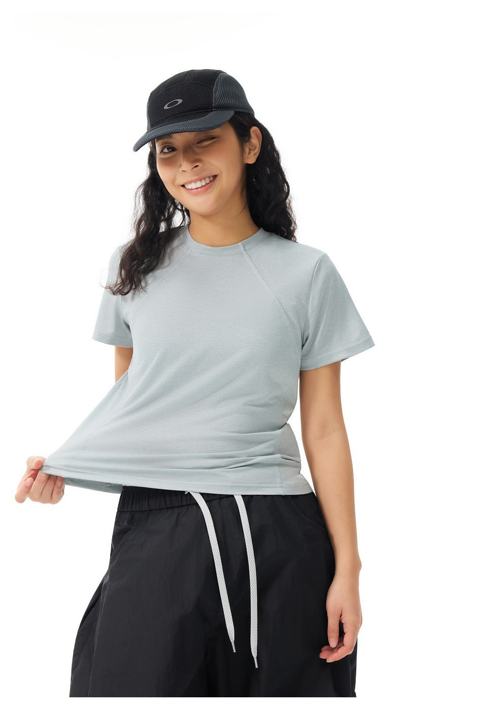 Model wearing a pale gray short-sleeve shirt and black pants, pulling the shirt hem outward to show its fit.