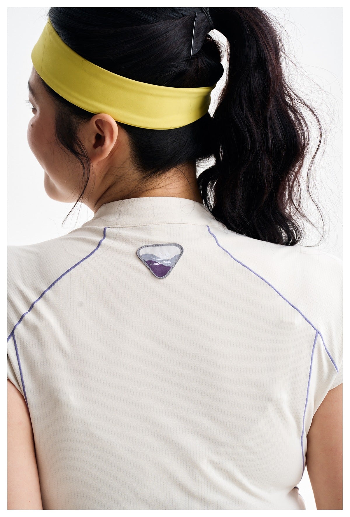 Close-up rear view of an ivory sports top with purple seam accents and a small triangular logo patch at the upper back.