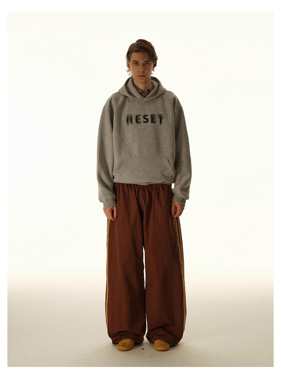 Model wearing a light gray hoodie with dark blurred chest lettering and wide rust-brown pants with side stripes.