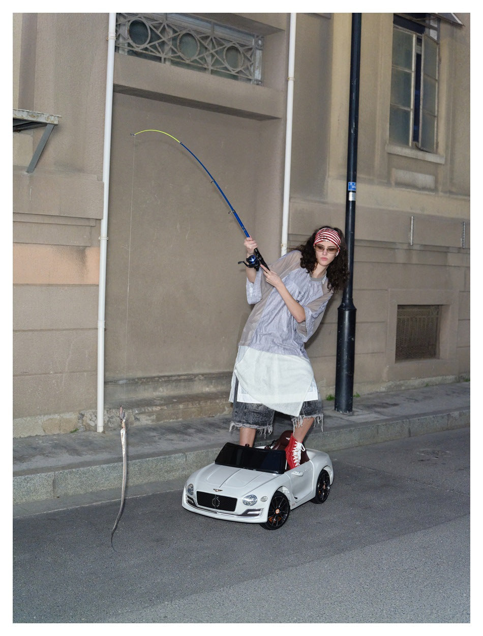 Model wearing the gray and taupe short-sleeve top over denim shorts, holding a fishing rod while standing in a toy car