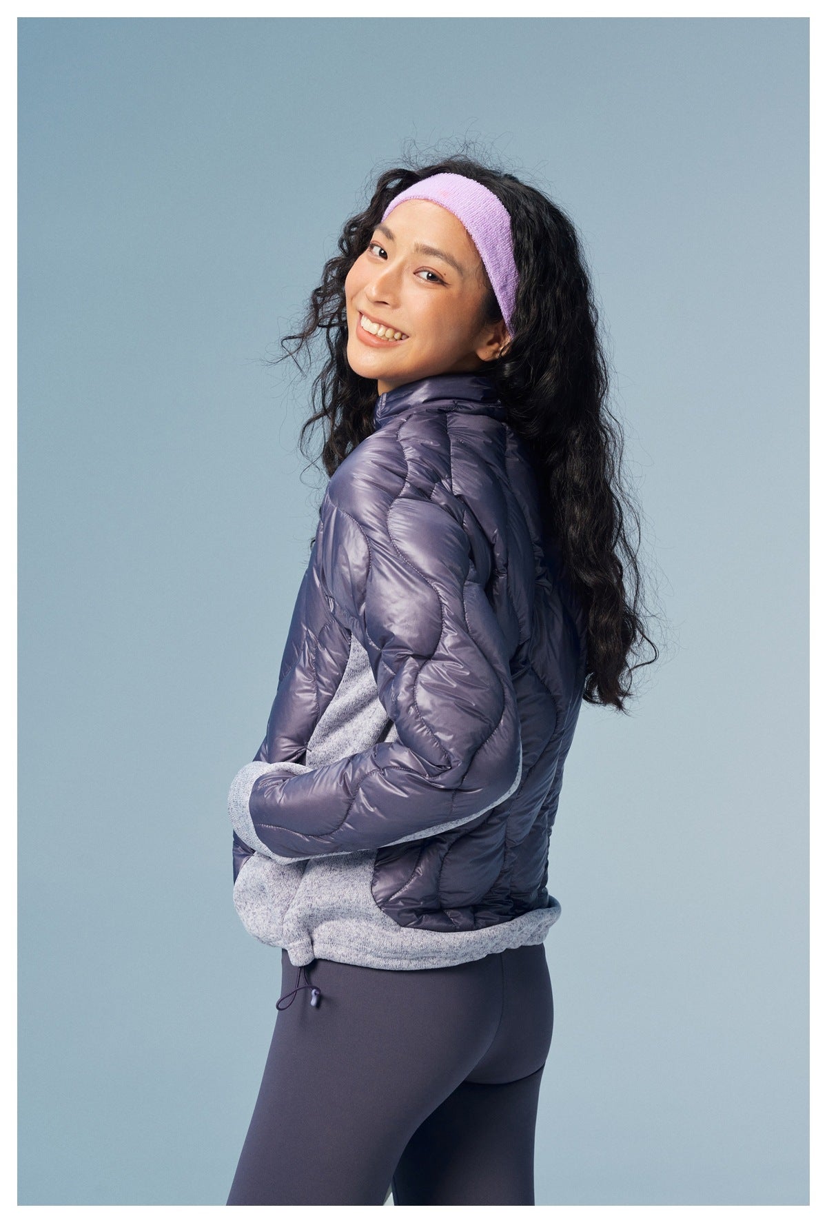 Model in a purple quilted jacket with gray panels, shown from the side against a blue background.